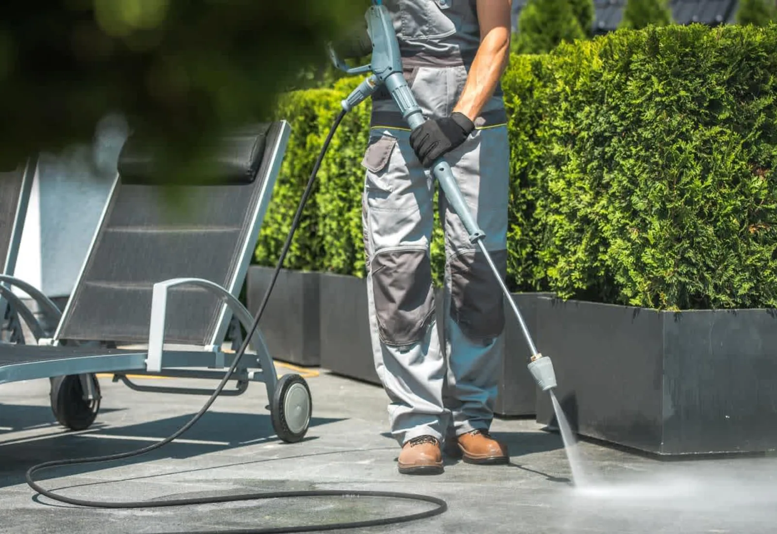 Worker pressure washing a backyard concrete patio with a spray wand next to lounge chairs