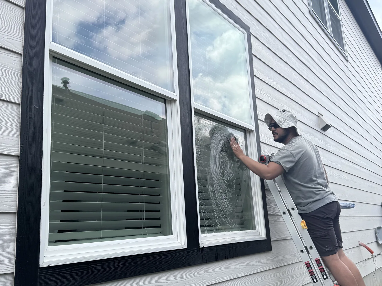 Technician on a ladder hand washing a home's window, soap swirled across the glass